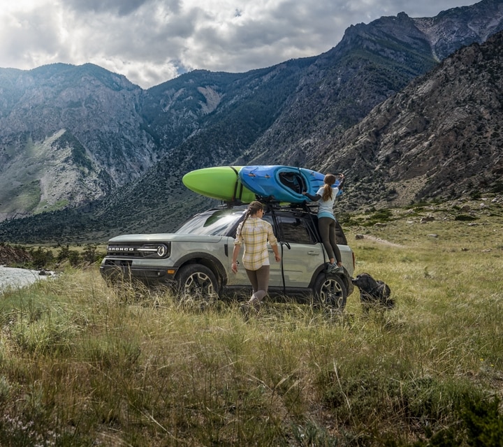 Two people are standing outside a 2024 Ford Bronco® Sport that is parked by a lake with a kayak on the roof