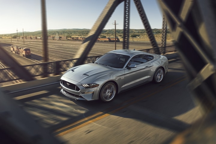 A 2023 Ford Mustang® GT model in Iconic Silver Metallic being driven across a metal bridge over a train yard