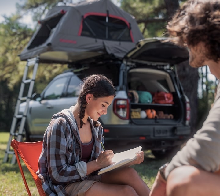 A 2024 Ford Bronco® Sport is in the background while a young woman writes in her journal and a man sits near her