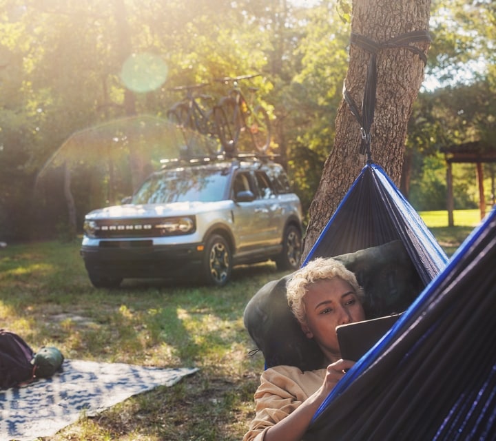 A lady lying in a hammock with gear on a blanket behind her with a 2024 Ford Bronco® Sport is in the background