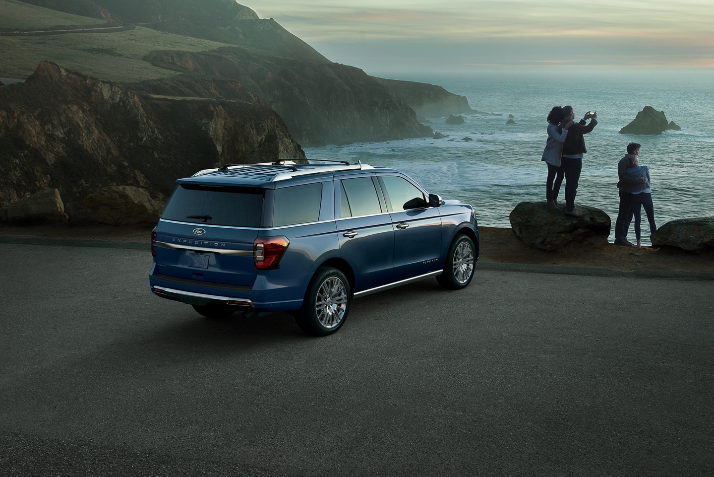 A group of people take a picture of the sunset at an ocean overlook while a Ford Expedition SUV is parked in the foreground. 