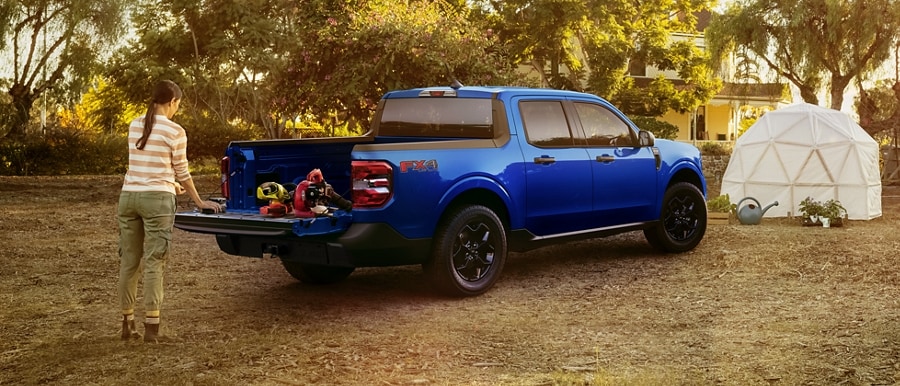 A 2024 Ford Maverick® truck in Atlas Blue Metallic is parked at a campsite with the bed being unloaded by a person