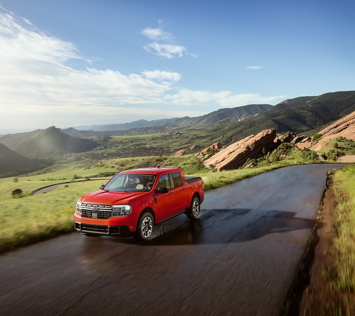 A 2024 Ford Maverick® truck in Hot Pepper Red is being driven on a road in a scenic location with mountains in the background
