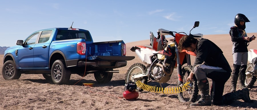 A 2024 Ford Ranger® in Velocity Blue is parked in the desert dunes next to two people with dirt bikes