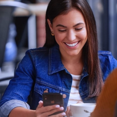 A woman sitting with a cup of coffee looking down at her smartphone and smiling.
