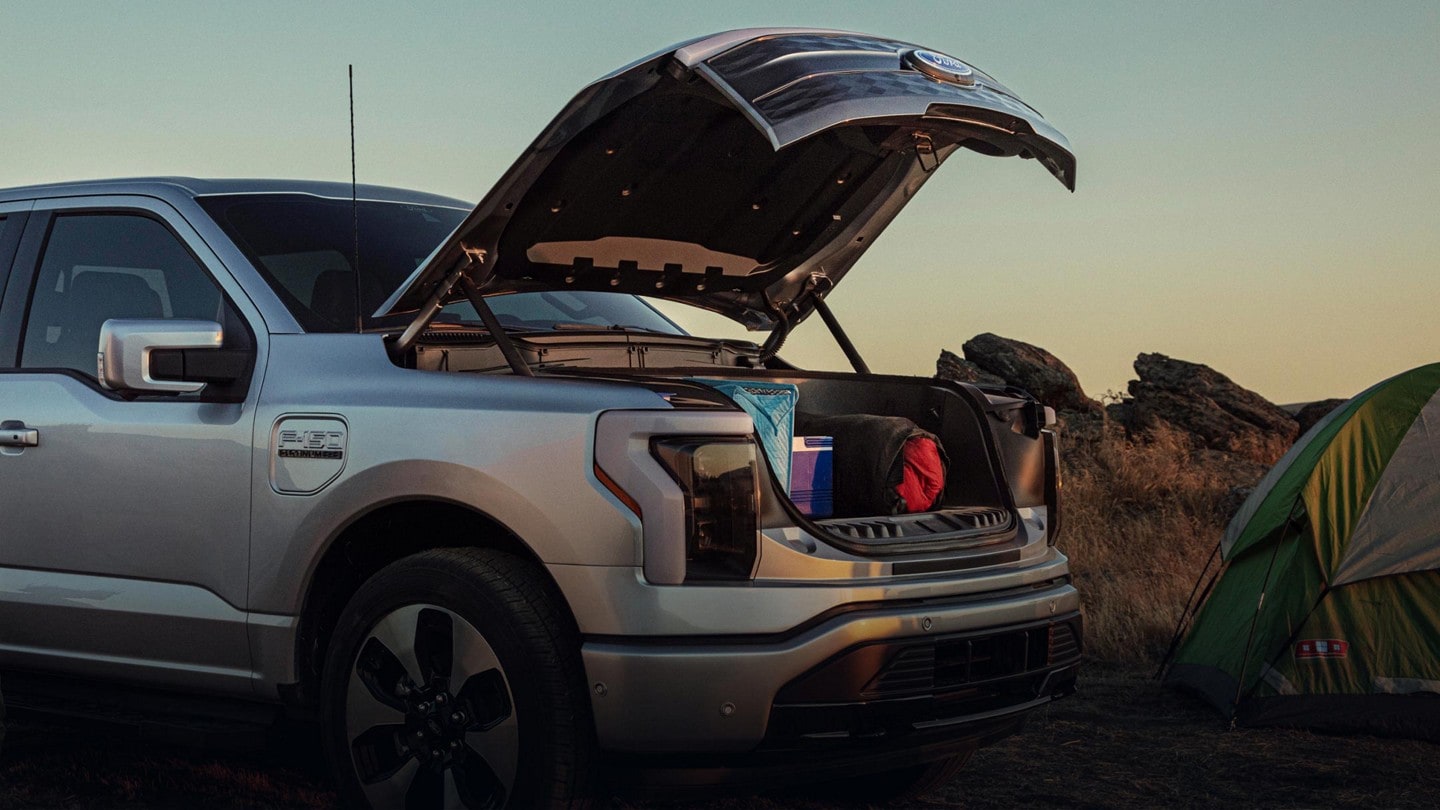 A silver 2023 Ford F-150® Lightning® parked at a campsite. Its FRUNK is open revealing camping gear inside