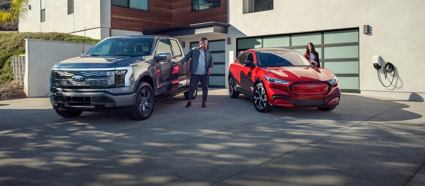 Two people open the driver doors of a gray 2023 Ford F-150 Lightning and red 2023 Ford Mustang Mach-E parked in the driveway