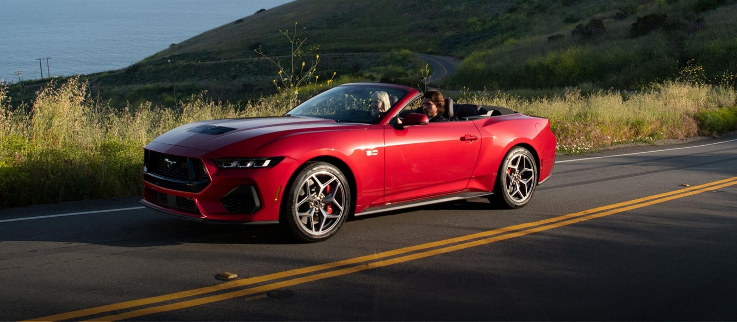 Two people in a red 2024 Ford Mustang convertible with its top down, driving on a seaside road at sunset