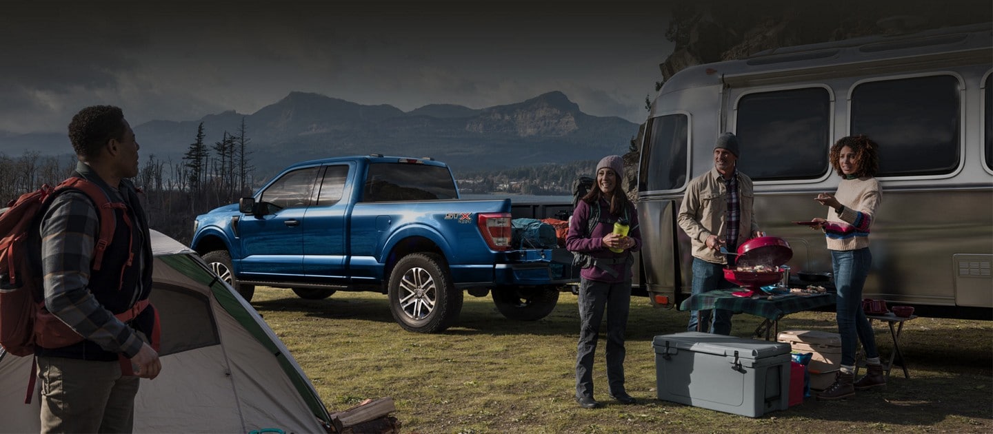 A 2022 Ford F-150 XL SuperCab 4x4 in Atlas Blue hooked up to an Airstream camper surrounded by people grilling