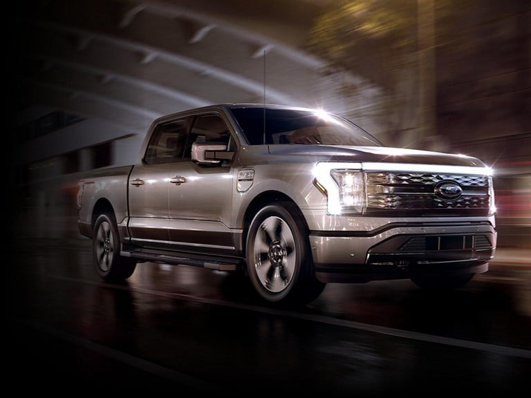 2023 Ford F-150® Lightning® in silver parked on a rain-slicked urban road at night with headlamps on