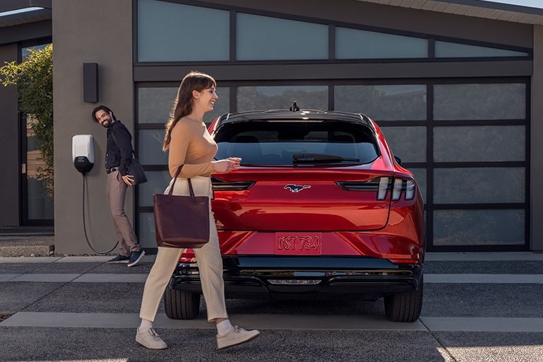 Woman in business casual attire walking around the rear of a 2023 Ford Mustang Mach-E® parked in the driveway as a man unplugs the vehicle