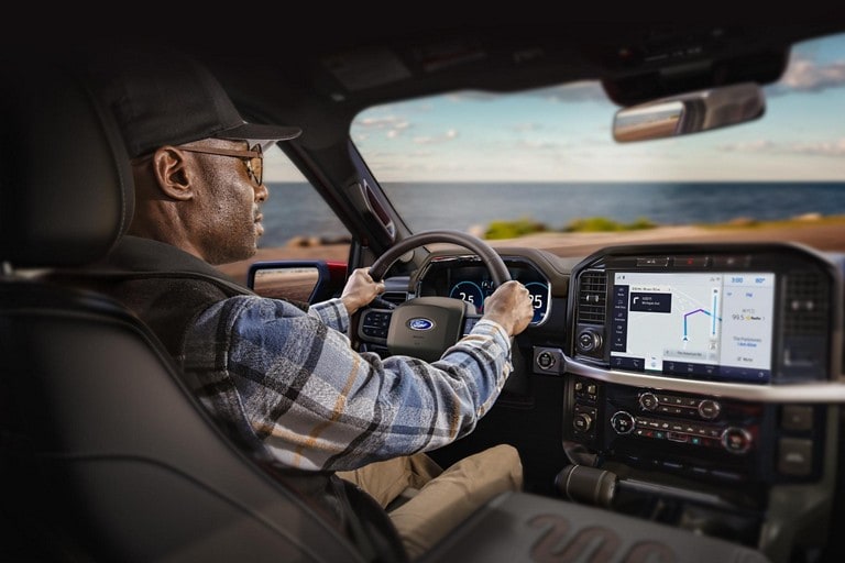 A person behind a wheel driving a 2022 Ford F-150 with the ocean in the background