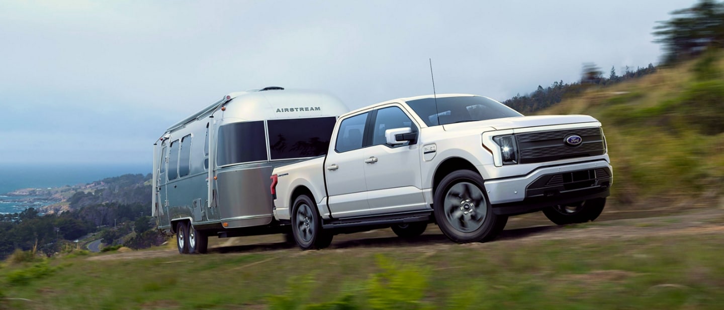 A white 2023 Ford F-150® Lightning® towing a silver camper up a hill
