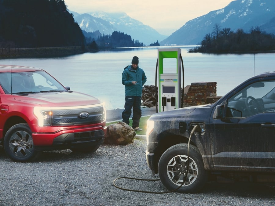 On the left, a red 2023 Ford F-150® Lightning®. On the right, a black one plugged into a public charger. A lake is in the distance