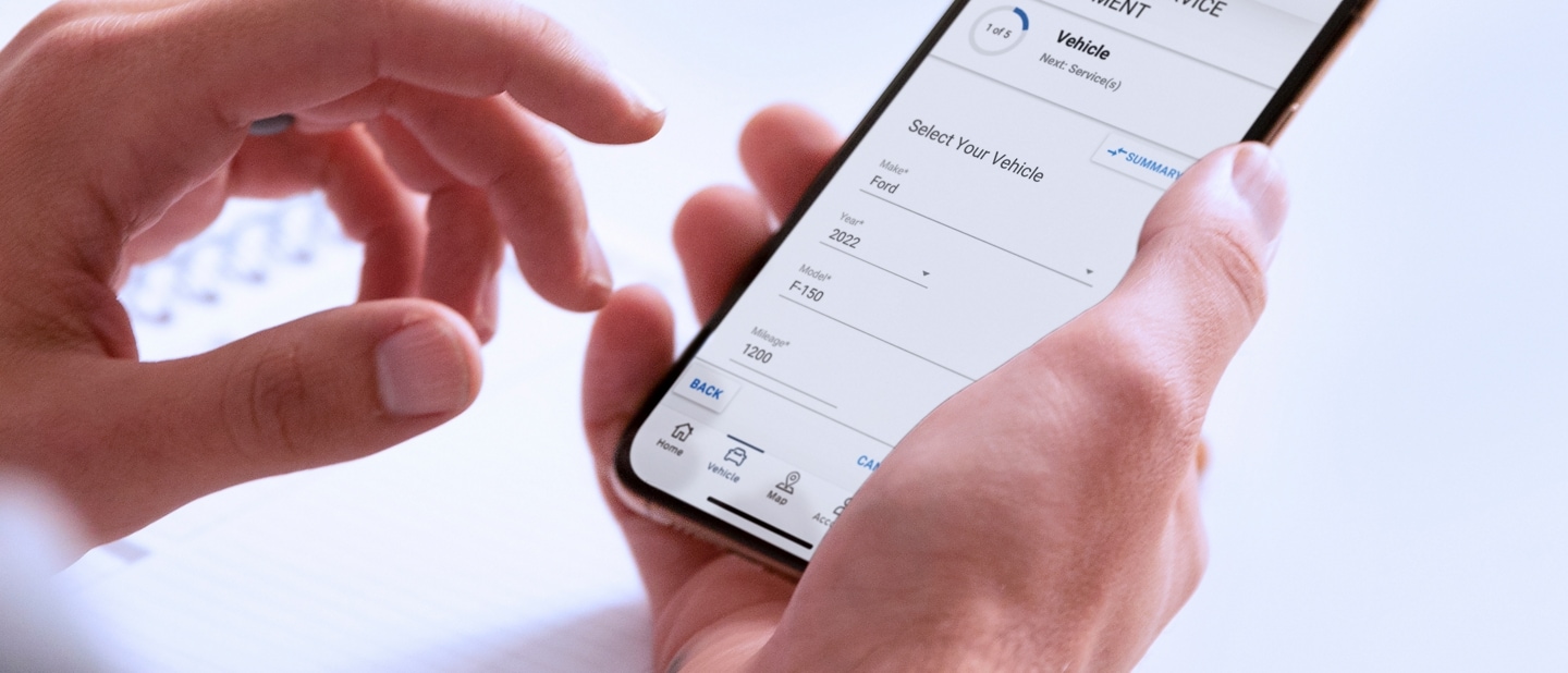 A hand holds a smartphone displaying the Schedule Service menu in the FordPass™ App