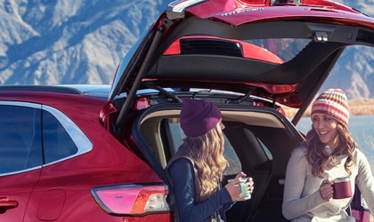 o	Two women sitting on the back of a 2022 Ford Escape® SUV parked by a body of water drinking coffee while a man is fishing nearby